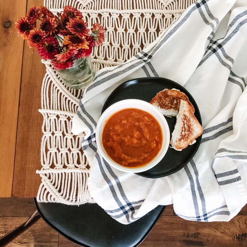 An image of a steamy bowl of tomato bisque soup, with herbed grilled cheese sandwich pieces on the side, sits at a well-dressed table.