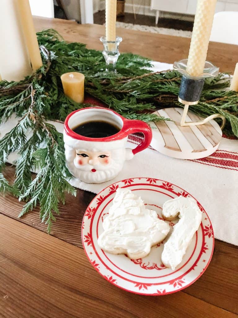 A festive holiday table with greenery and candles, adorned with an adorable Santa mug full of coffee next to a plate with two sugar cookies with buttercream icing.