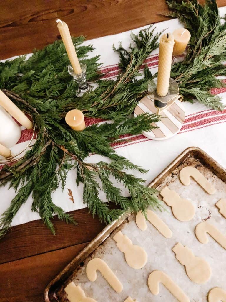 Freshly cut sugar cookie dough cut-outs sit on a baking pan on top of a table adorned with fresh holiday greenery and candles.