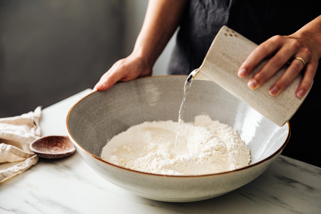 Midsection of woman pouring water on wheat flour in bowl.