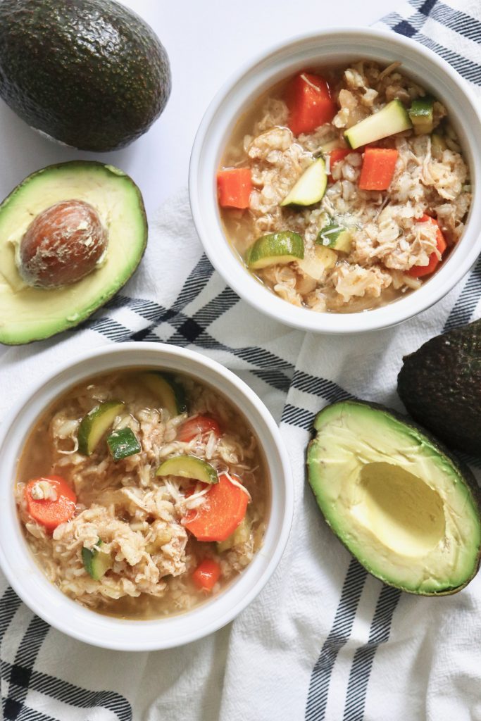 An image of two bowls of chicken tortilla soup sitting on a tablecloth-laden table, surrounded by cut avocados.