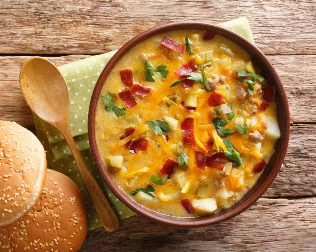 Creamy cheeseburger soup close ups with chives and bacon on a wooden table with a green napkin, wooden spoon, and burger bun