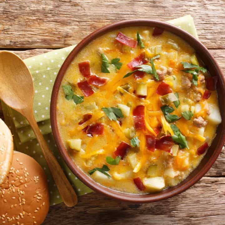 Creamy cheeseburger soup close ups with chives and bacon on a wooden table with a green napkin, wooden spoon, and burger bun