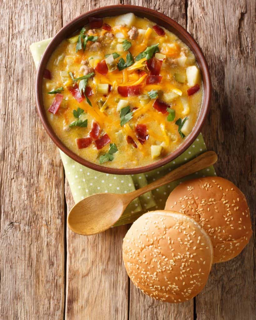 Brown bowl of delicious looking cheeseburger soup with chives and bacon on a green napkin with a wooden spoon and burger buns