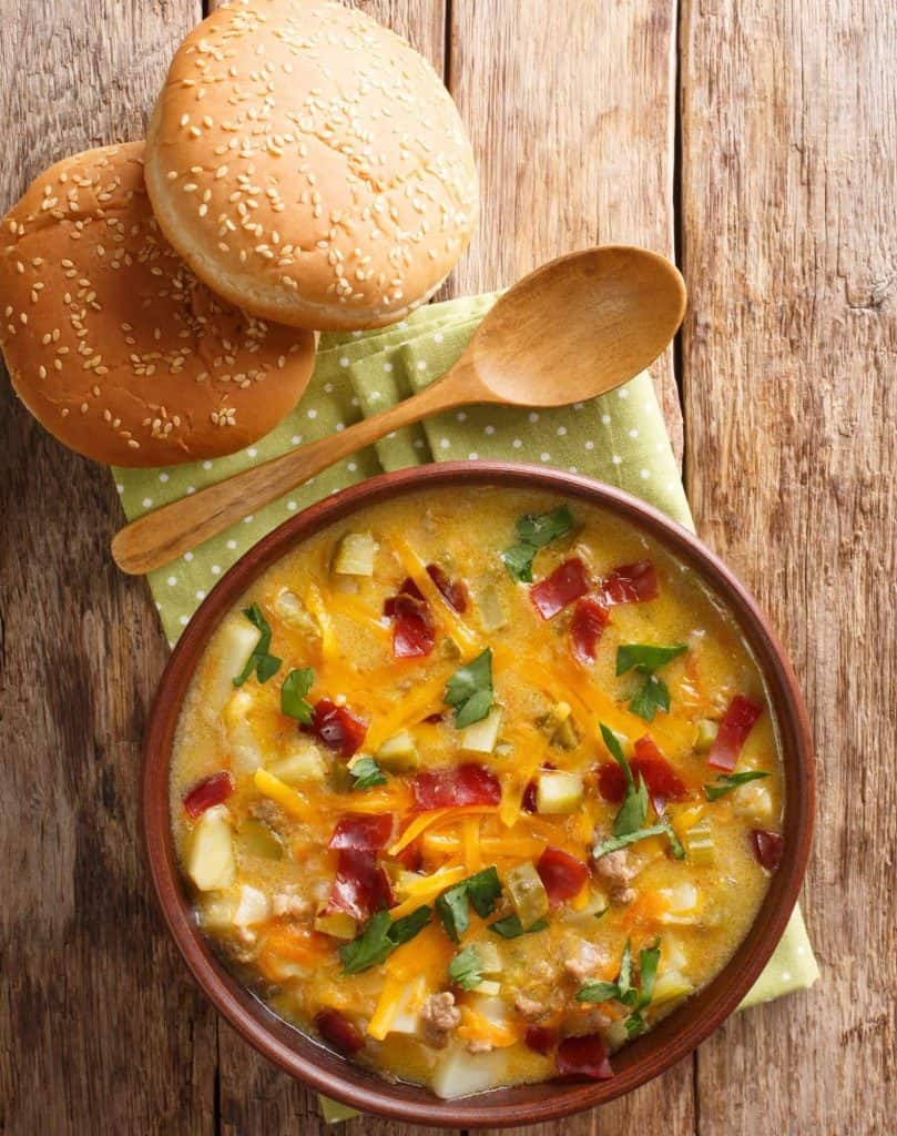 Close up of cheeseburger soup in a brown bowl topped with chives and bacon, placed with a spoon and burger buns.