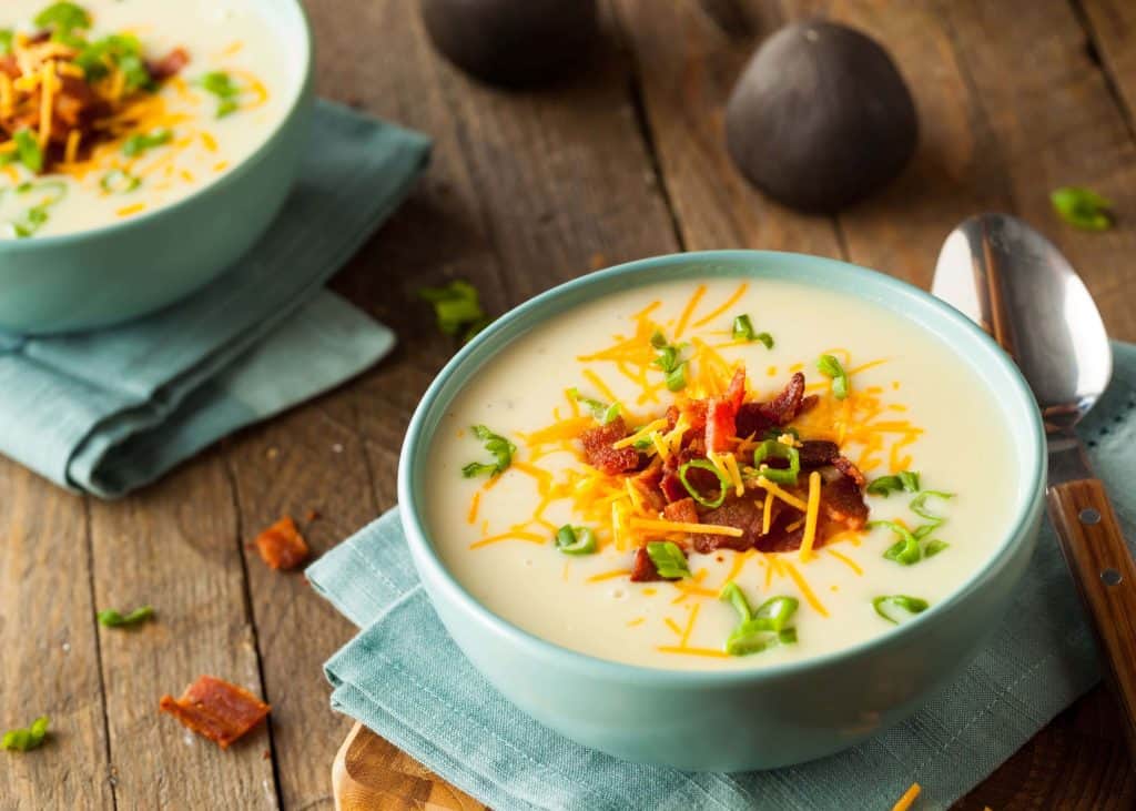 Two blue bowls of loaded potato soup with cheese, bacon and green onions on a wooden table