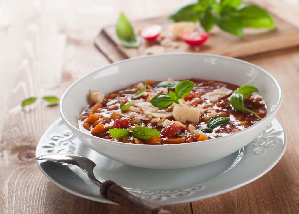 Minestrone soup garnished with basil and parmesan in a while bowl on a wooden table