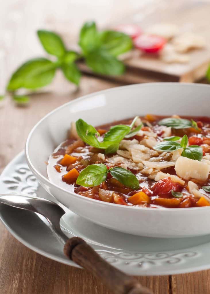 Close up of Minestrone soup garnished with basil and parmesan in a while bowl on a wooden table