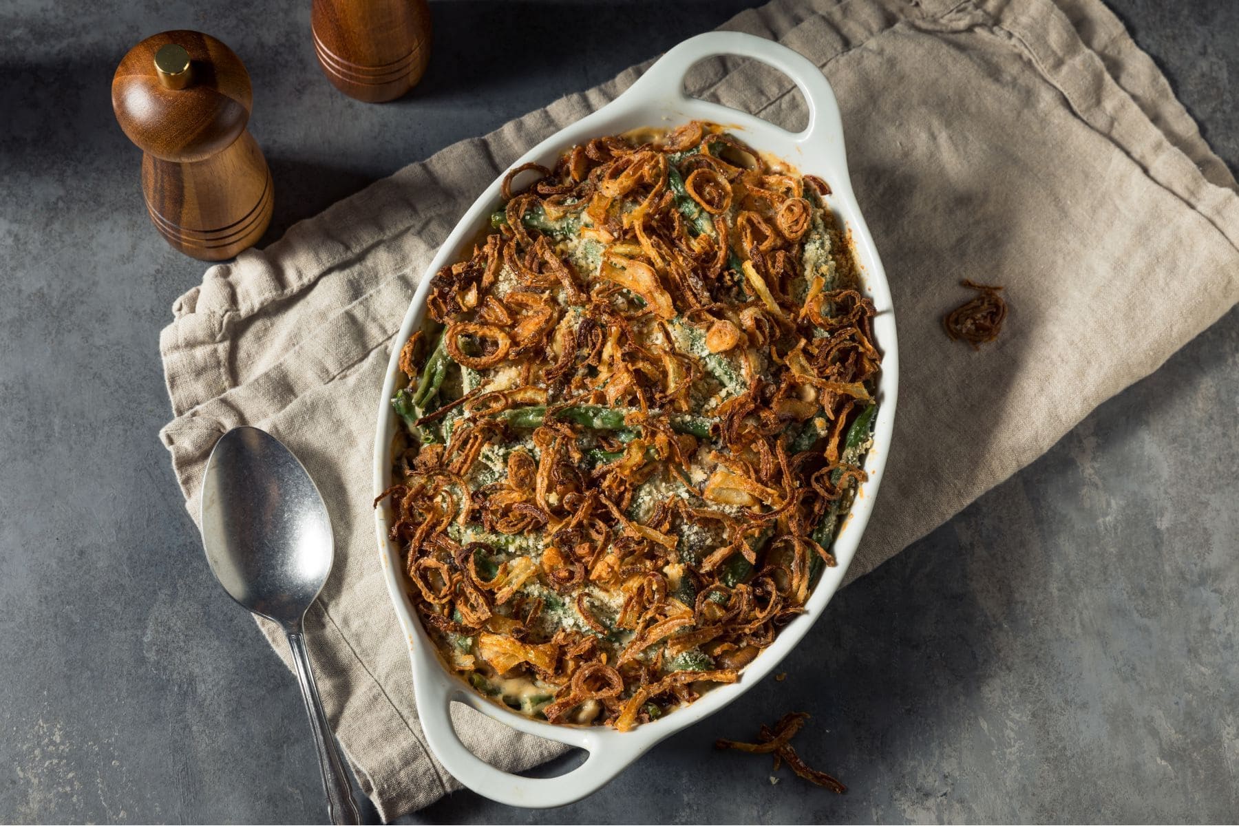 A green bean casserole in a white ceramic baking dish topped with homemade onion crisps sitting atop a beige tea towel on a grey table.