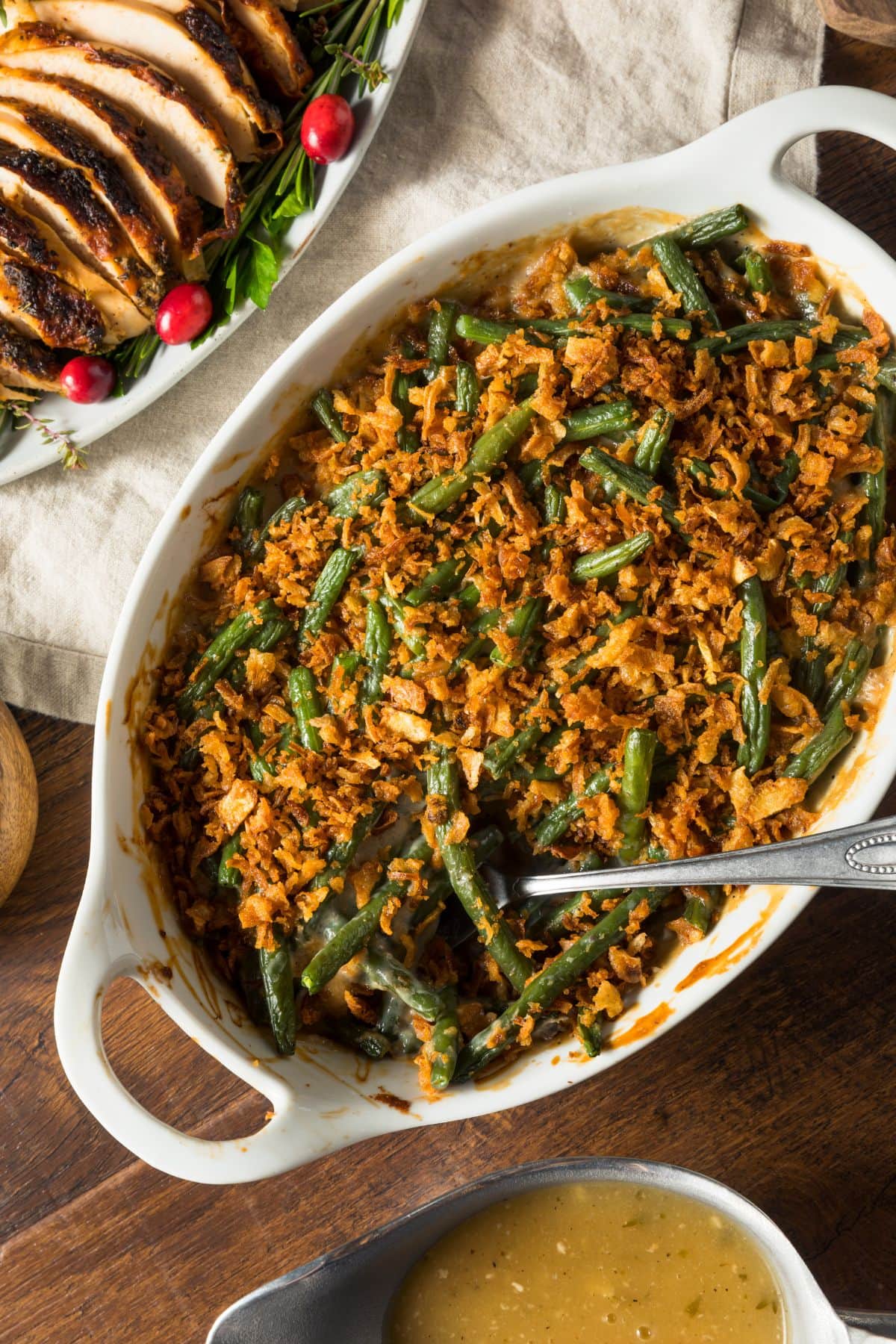 A healthy, homemade green bean casserole in a white dish beside a tray of sliced turkey and gravy on a wooden table.