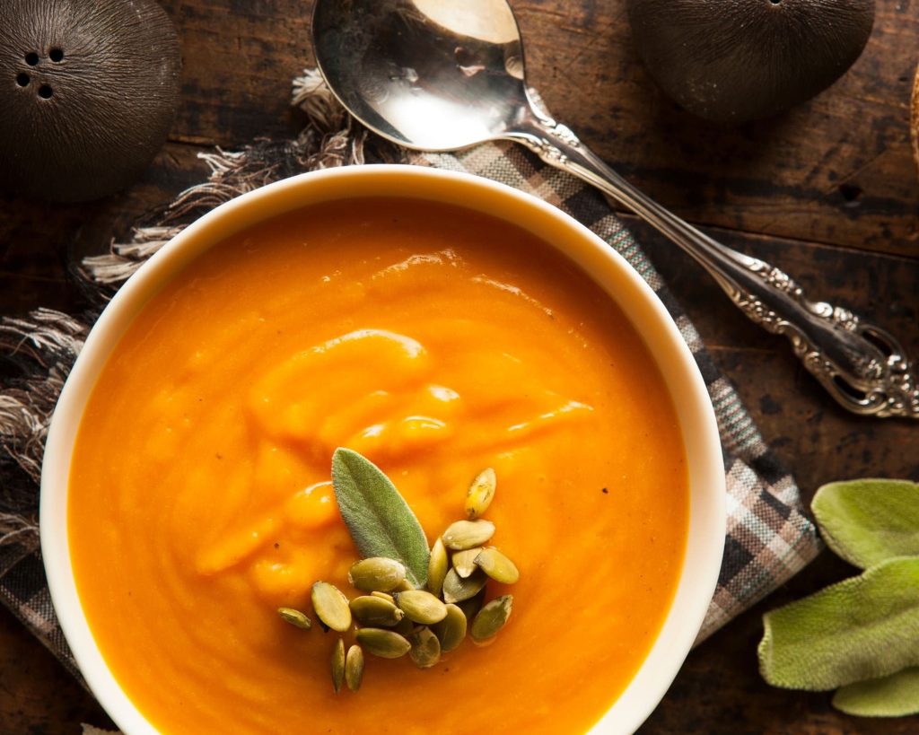 A bowl of warm, delicious butternut squash carrot soup topped with fresh pumpkin seeds and surrounded by sage, a spoon, and a napkin on a wooden table - the perfect autumn meal!