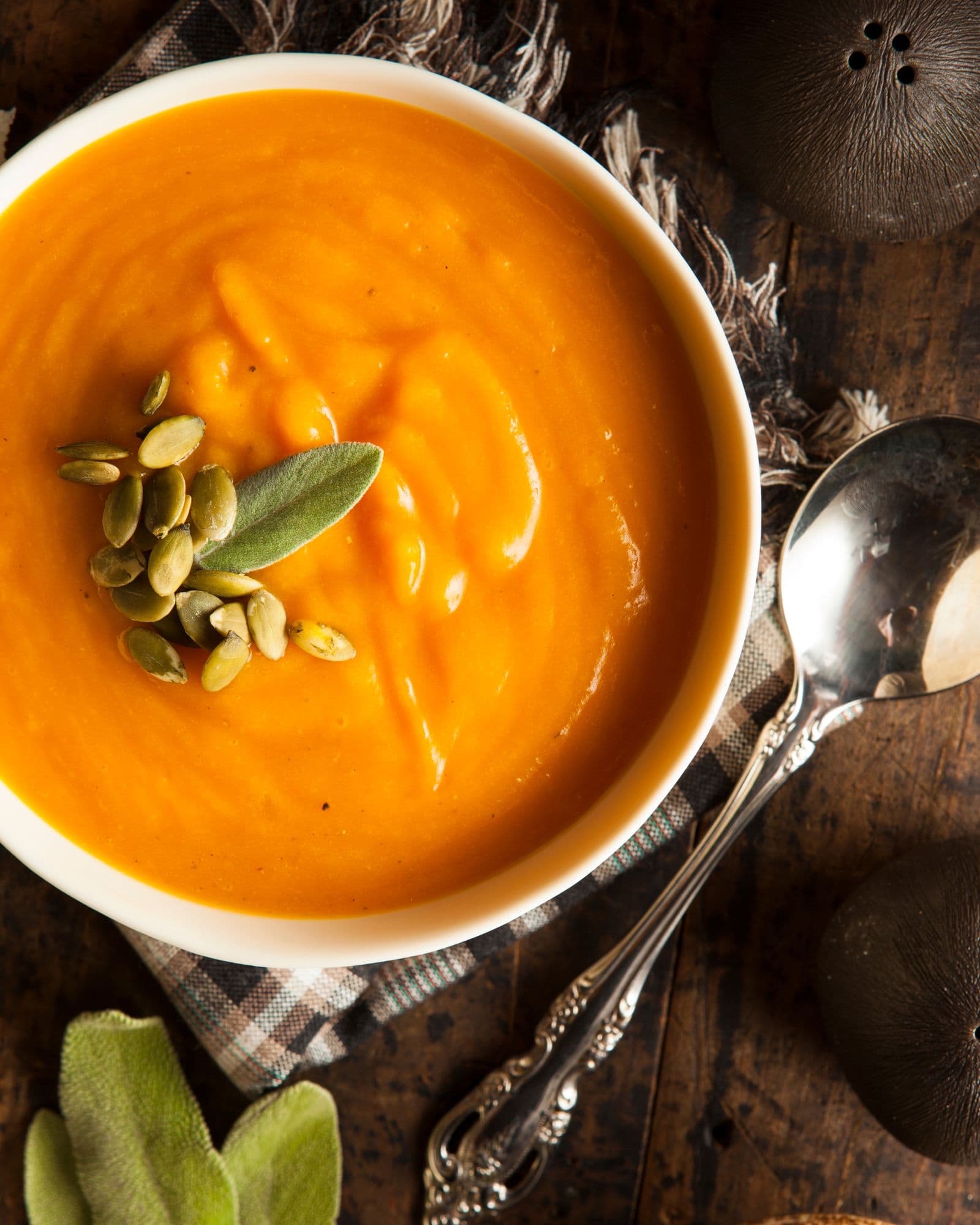 Bowl of creamy butternut squash carrot soup beside fresh sage, a spoon, and a checkered napkin on a wooden cutting board.