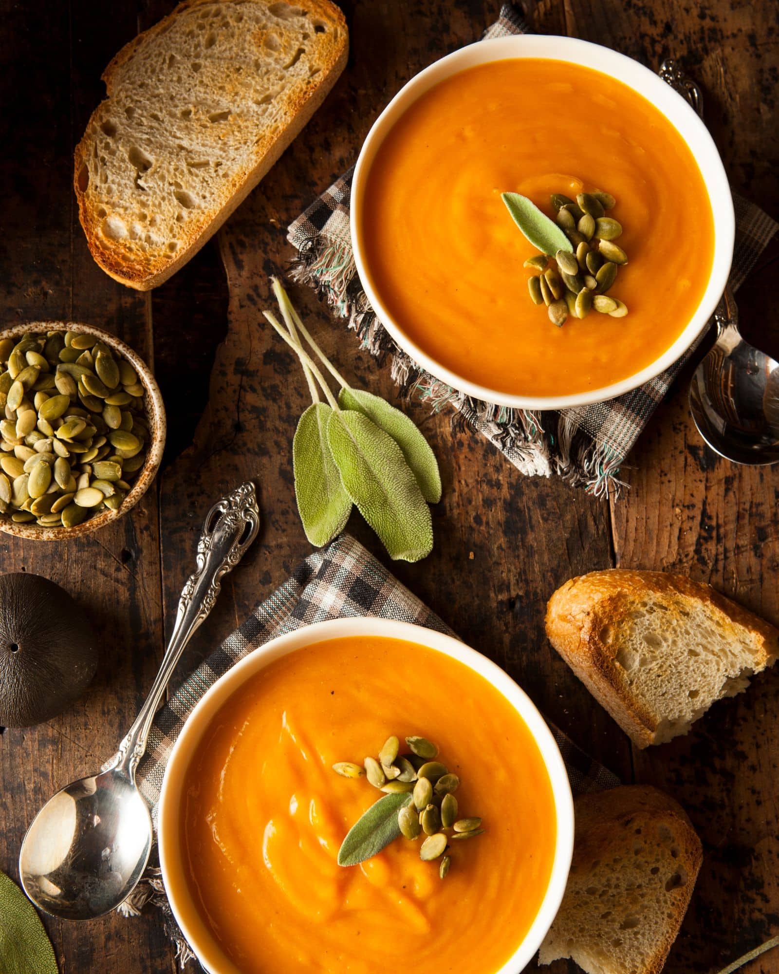 Two bowls of warm, delicious butternut squash carrot soup topped with fresh pumpkin seeds and surrounded by sage, fresh crusty bread, spoons, and napkins on a wooden table - the perfect autumn meal!