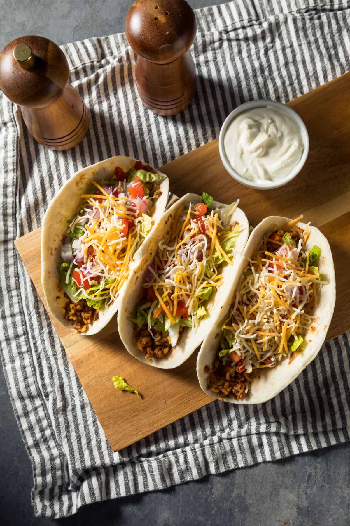 sourdough tortillas filled with delicious taco ingredients--ground meat, lettuce, onions, tomatoes, and topped with cheese--sitting on a cutting board with sour cream.