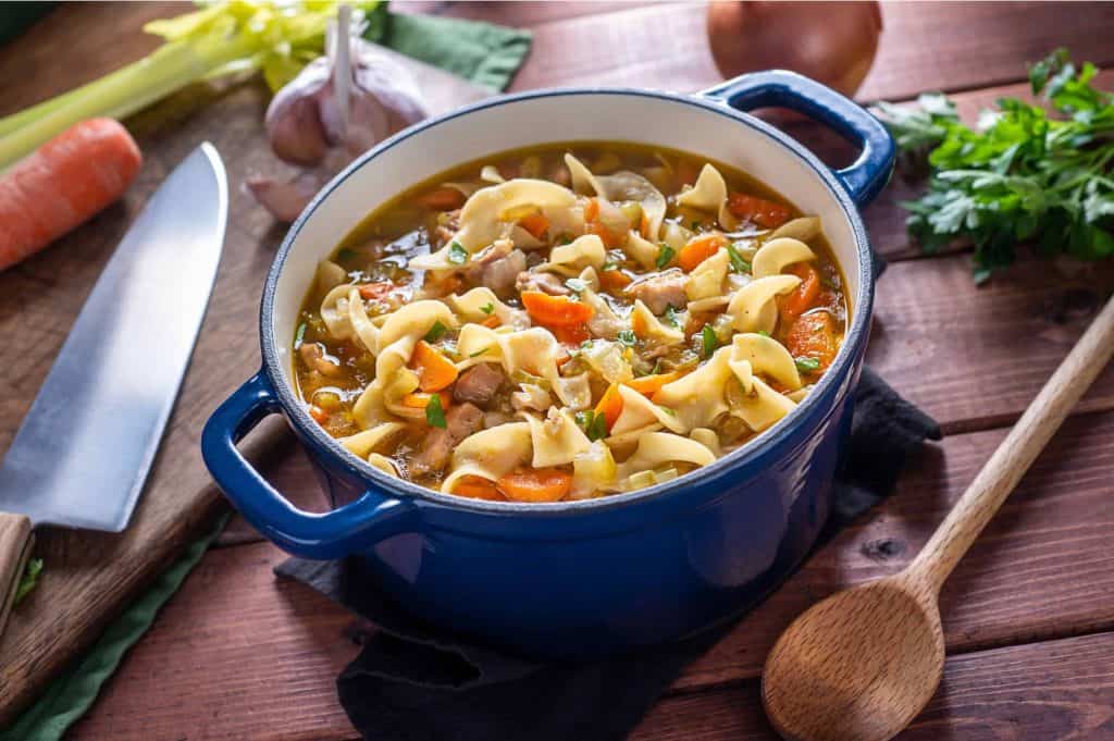 Close up of gluten-free chicken noodle soup in a green dutch oven surrounded but garlic, onion, a raw carrot, and a knife.