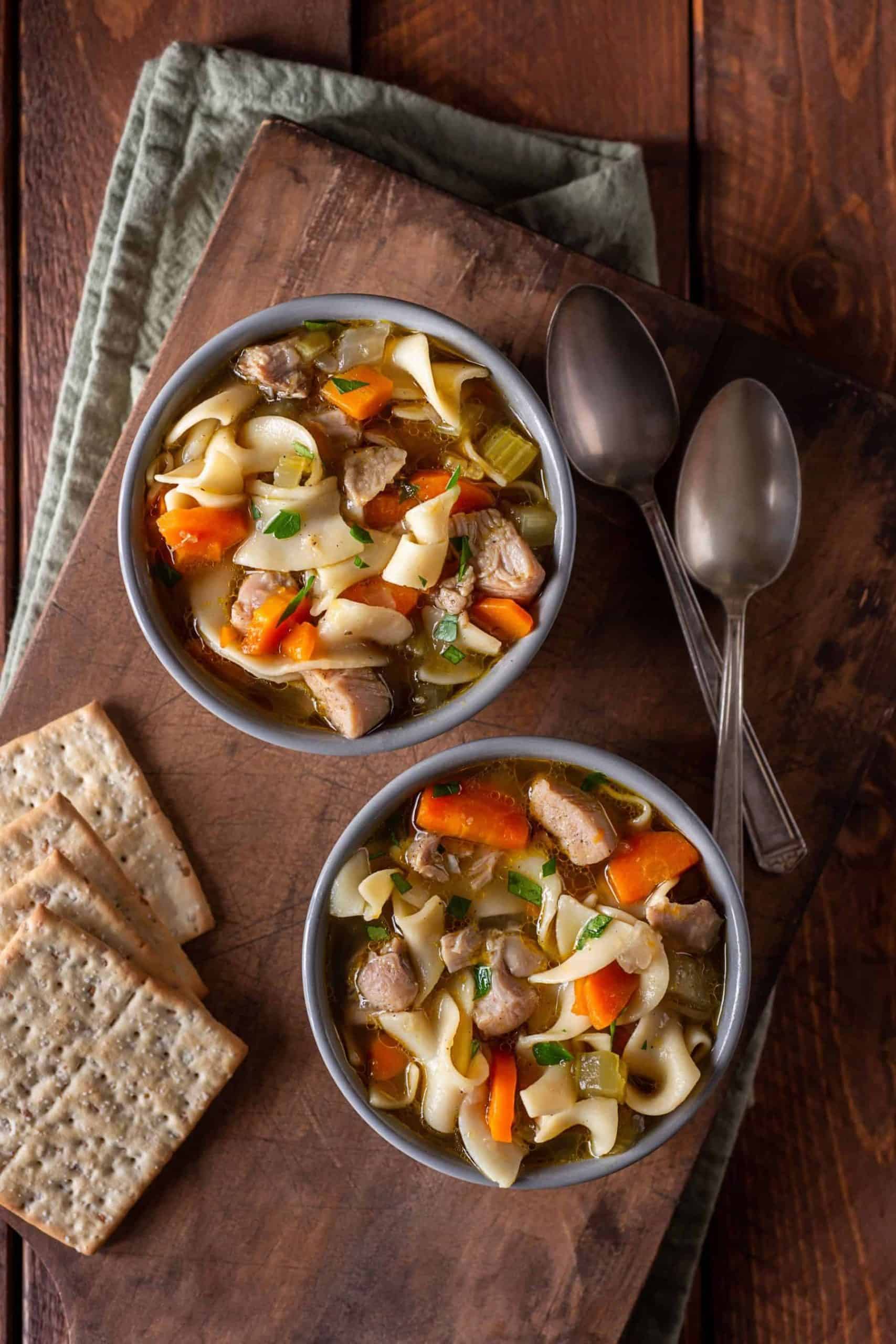 Two bowls of gluten-free chicken noodle soup on a wooden cutting board with crackers and spoons.