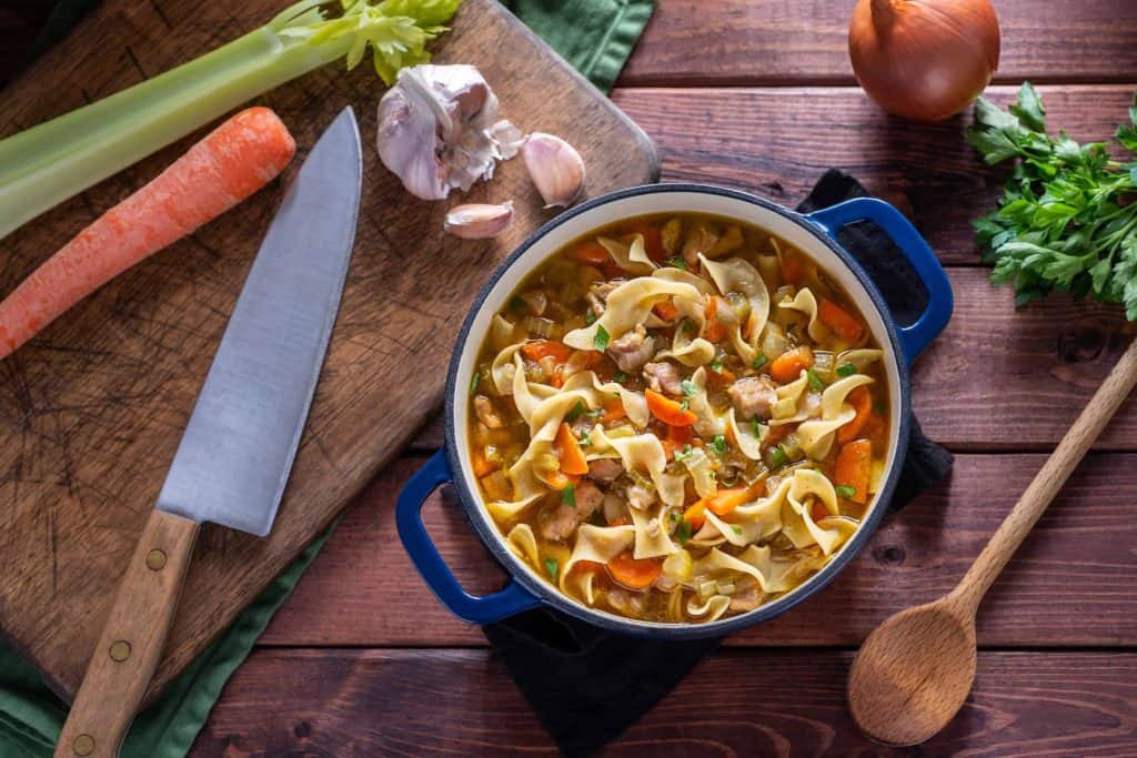 Close up of chicken noodle soup in a green dutch oven surrounded but garlic, onion, a raw carrot, celery, and a knife.