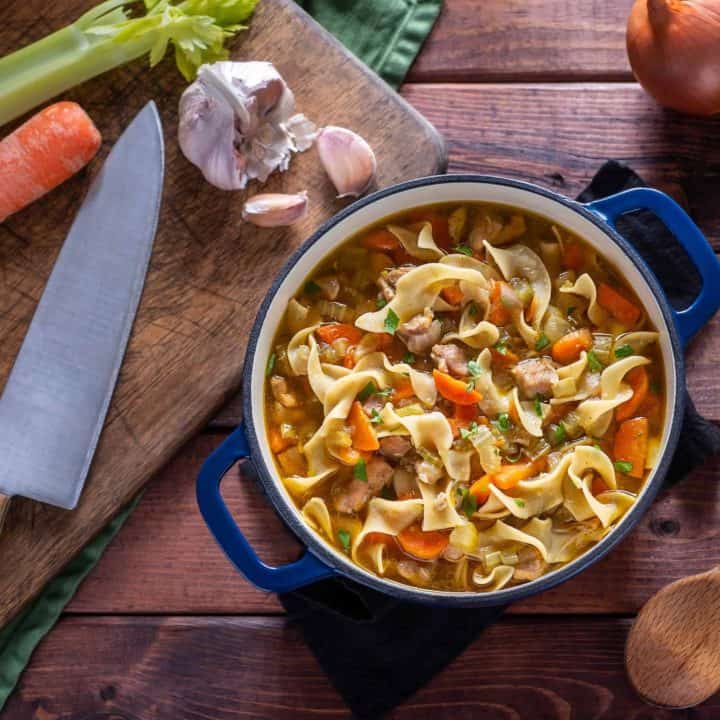 Close up of gluten-free chicken noodle soup in a green dutch oven surrounded but garlic, onion, a raw carrot, and a knife.