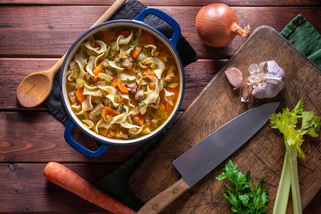 Close up of gluten-free chicken noodle soup in a green dutch oven surrounded but garlic, onion, a raw carrot, and a knife.