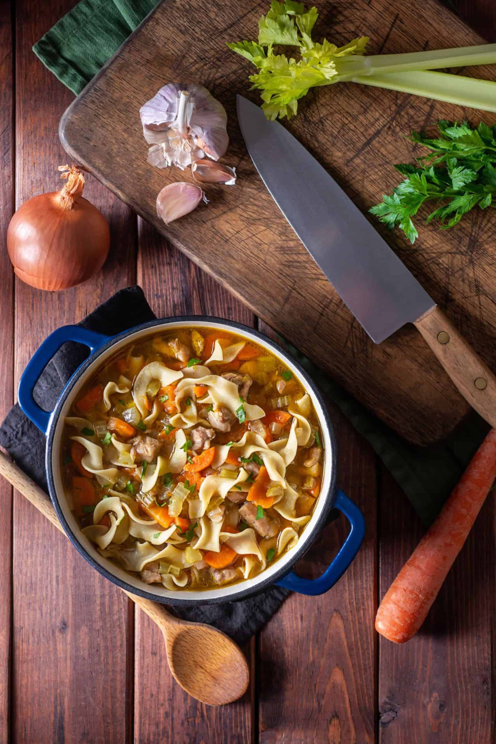 Garlic, celery, and a knife on a cutting board with an onion and carrot beside it, plus a dutch oven full of gluten-free chicken noodle soup