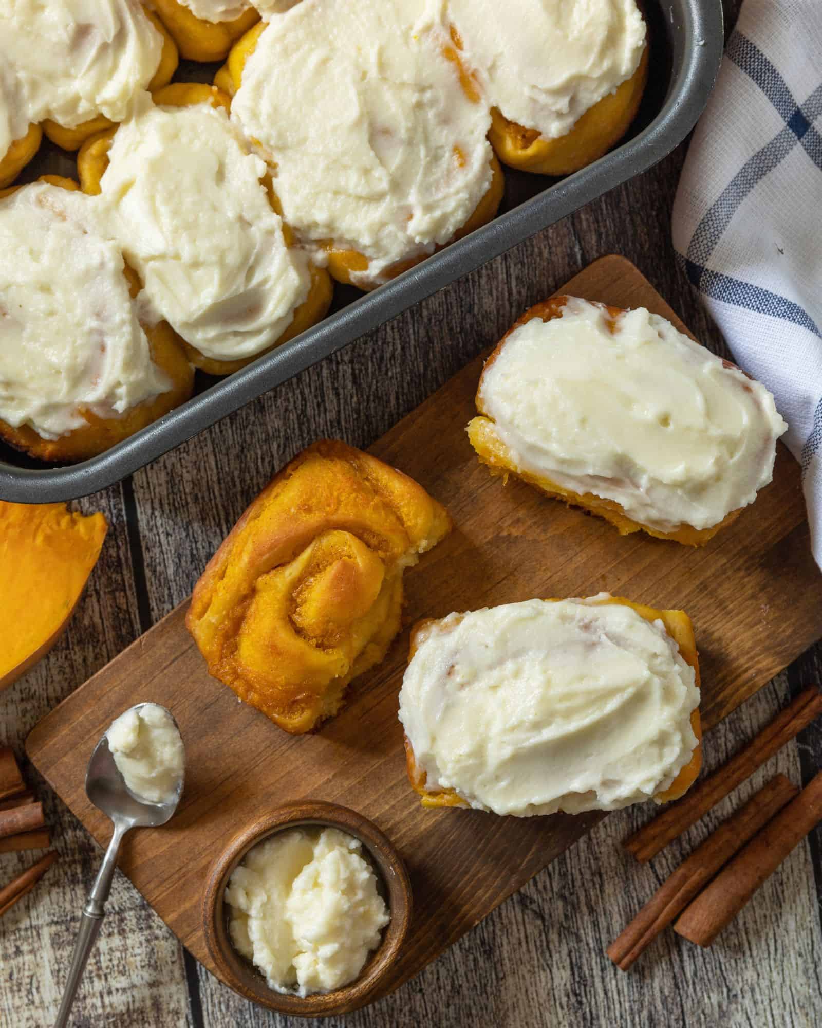 Immaculate pumpkin spice cinnamon rolls frosted in a pan beside cinnamon rolls on a wooden cutting board, waiting to be devoured