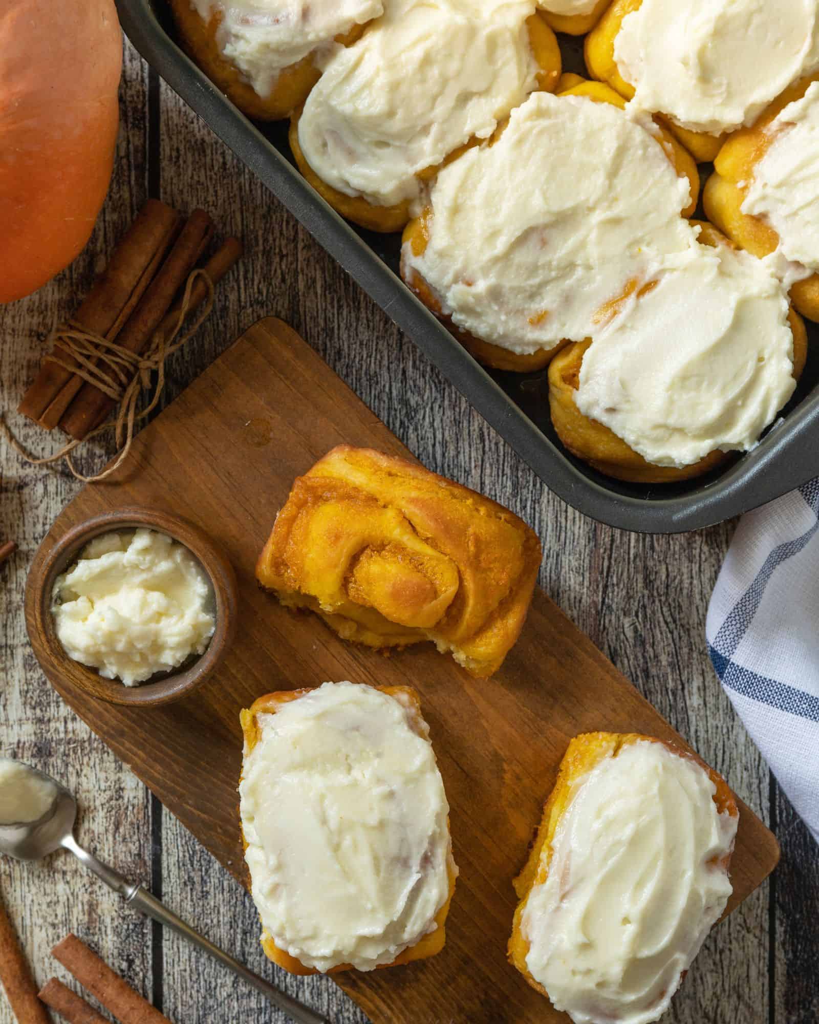 Immaculate pumpkin spice cinnamon rolls frosted in a pan beside cinnamon rolls on a wooden cutting board, waiting to be devoured