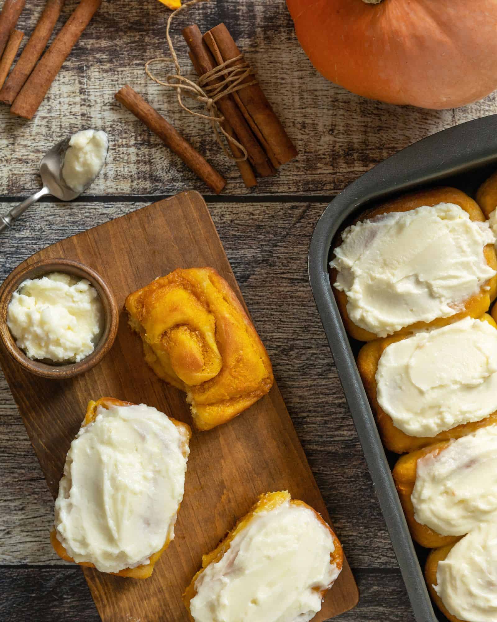 Warm, delicious pumpkin spice cinnamon rolls in a baking dish, topped with icing sitting beside a wooden cutting board with other cinnamon rolls, icing, cinnamon sticks, and pumpkins on a wooden table.