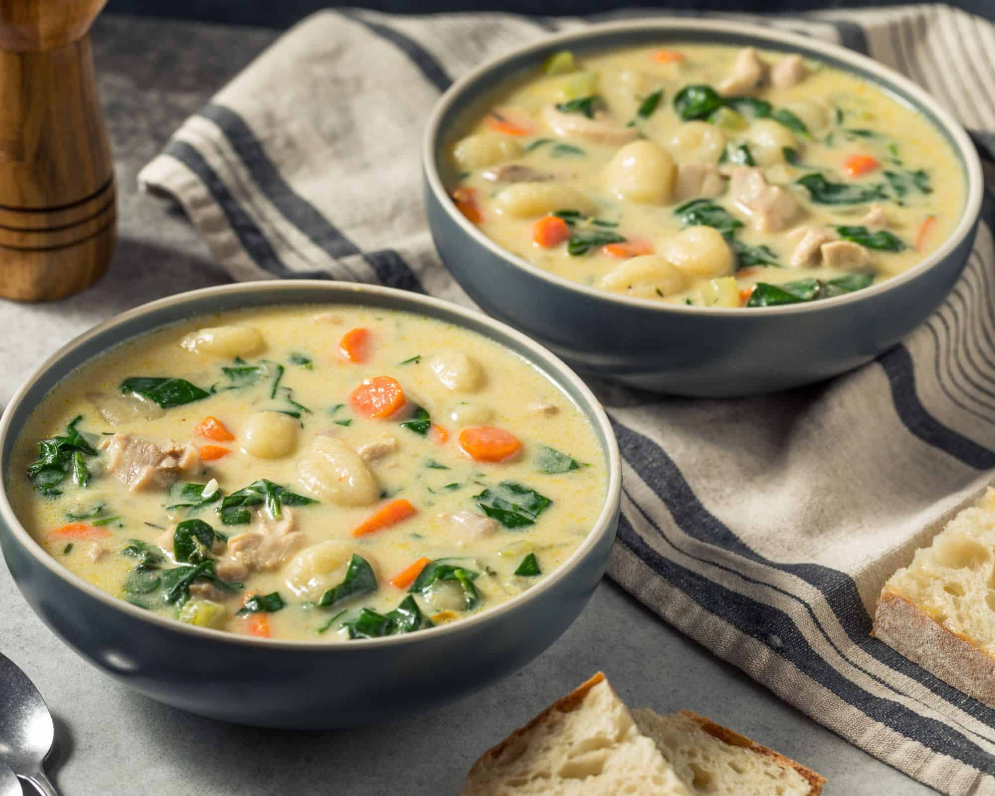 Two bowls of delicious chicken and gnocchi soup in dark grey bowls on a tea towel with fresh bread