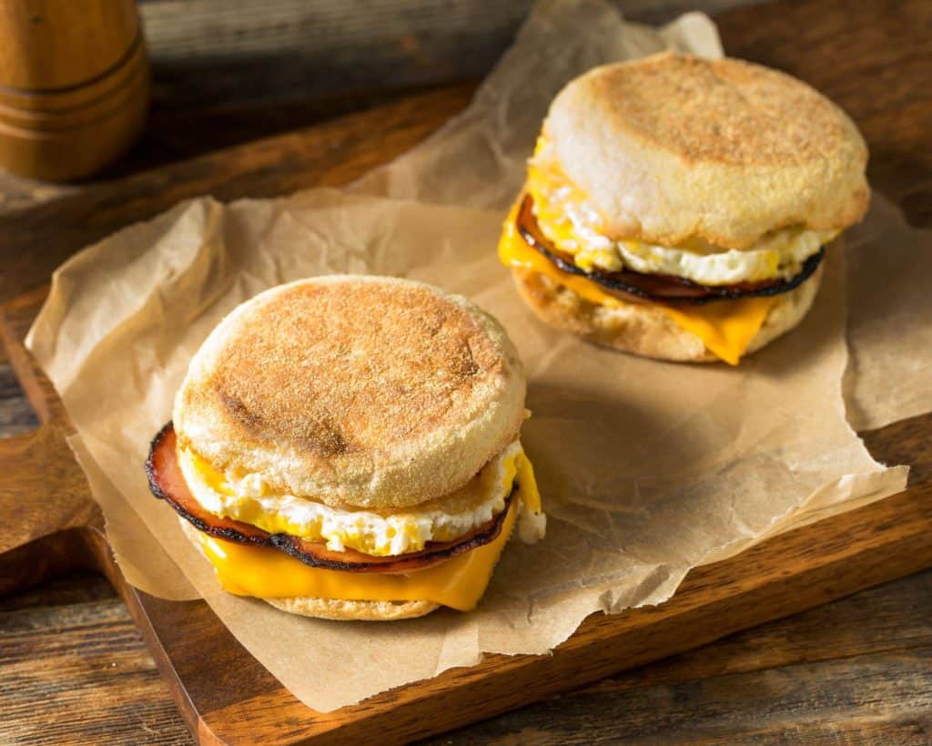 Two delicious English muffin breakfast sandwiches with egg, Canadian bacon, and melted cheese on a wooden cutting board.