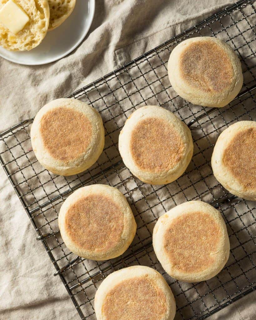 Fresh sourdough English muffins on a cooling rack with a beige towel and freshly sliced English muffin with butter and lots of nooks and crannies.