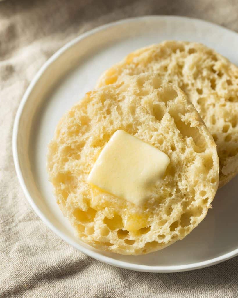 Fresh sourdough English muffin, sliced on a white plate with butter and lots of nooks and crannies.