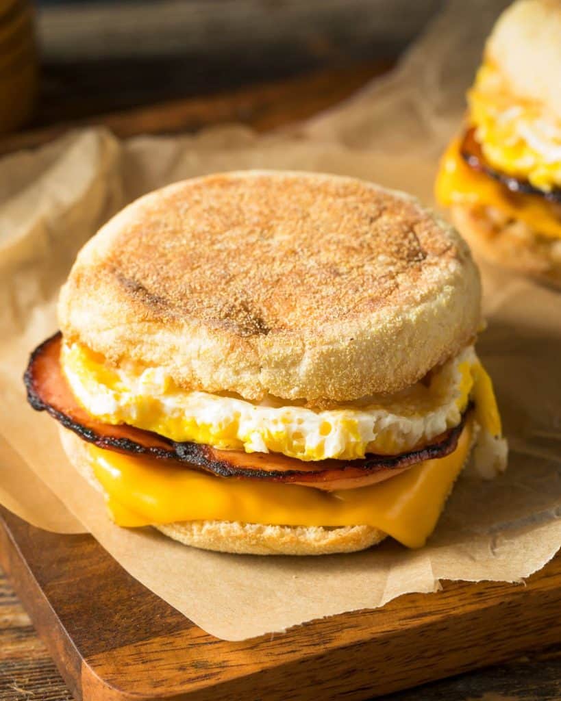 A delicious English muffin breakfast sandwich with egg, Canadian bacon, and melted cheese on a wooden cutting board.