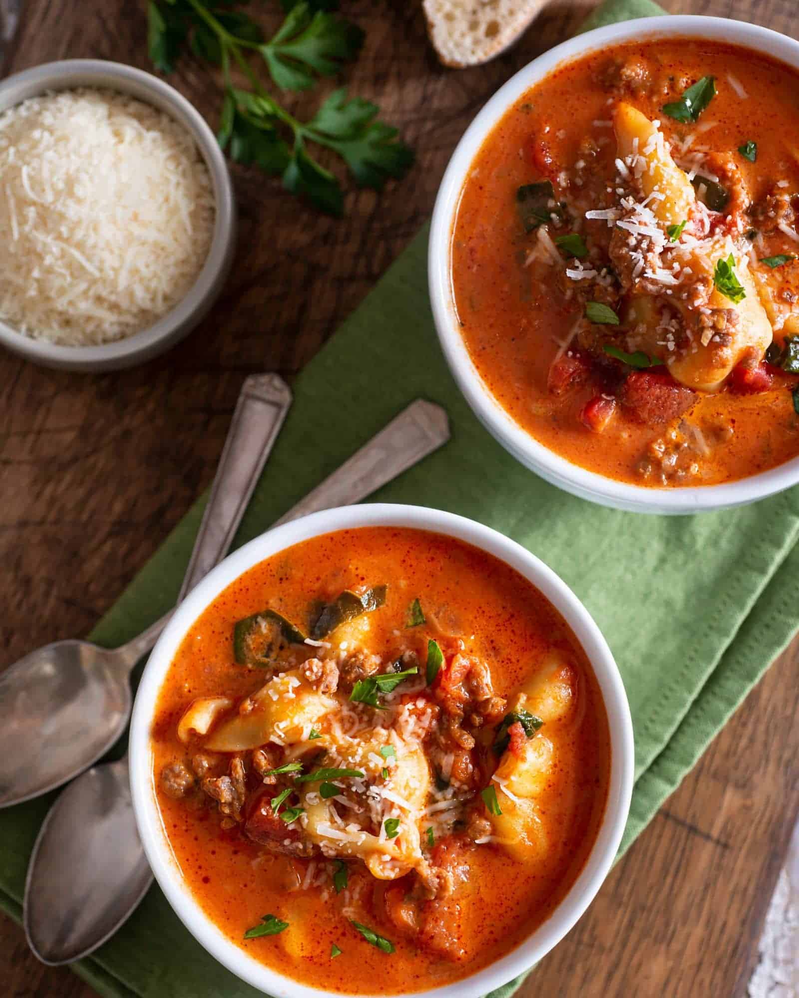 Two bowls of delicious, hearty tortellini soup with kale and ground beef on a cutting board with freshly grated parmesan and crusty bread.