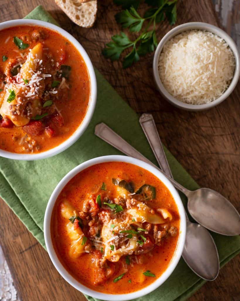 Two bowls of delicious, hearty tortellini soup with kale and ground beef on a cutting board with freshly grated parmesan and crusty bread.