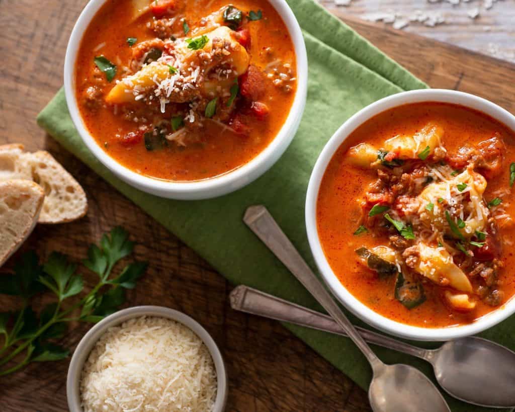 Two bowls of delicious, hearty tortellini soup with kale and ground beef on a cutting board with freshly grated parmesan and crusty bread.