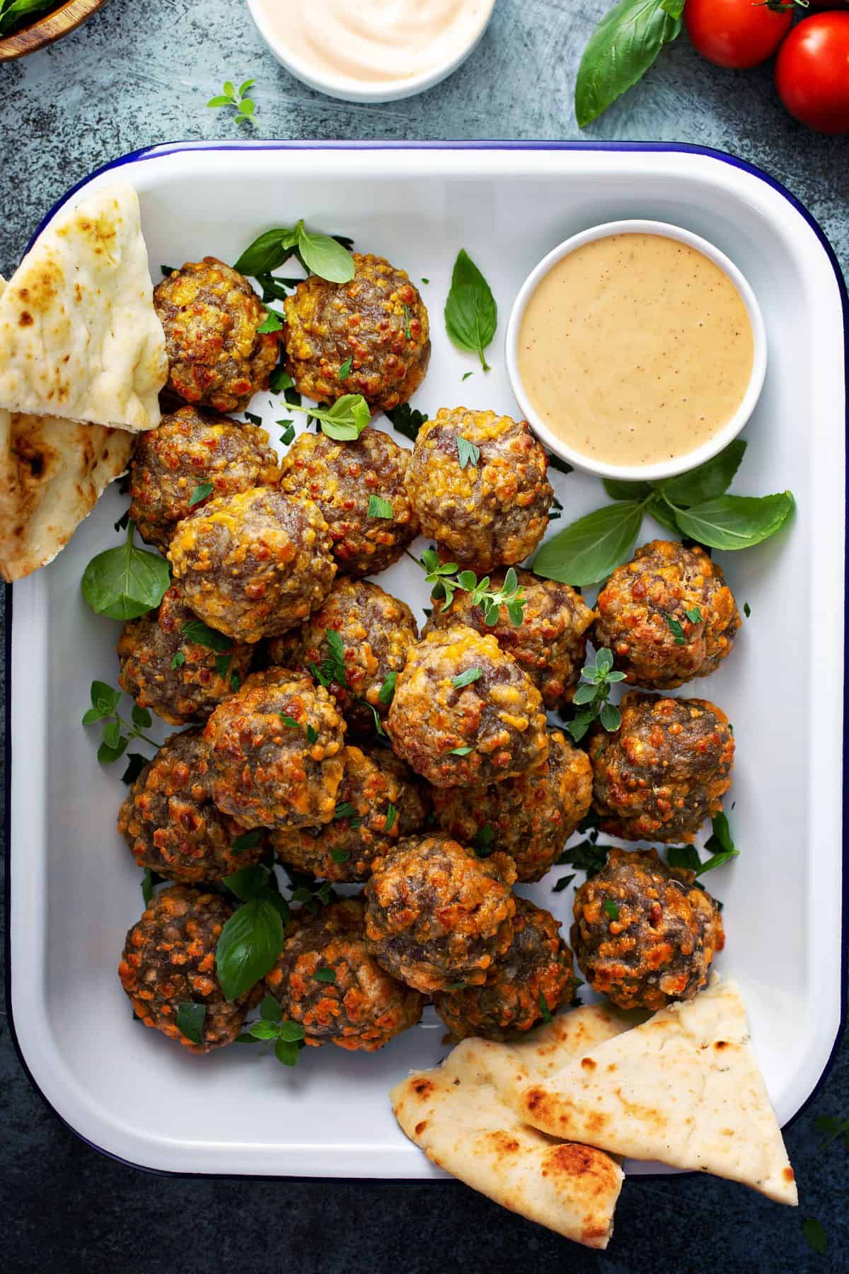 A white tray full of warm, delicious sausage balls surrounded by small bowls of dipping sauce and fresh pita bread  and fresh herbs.