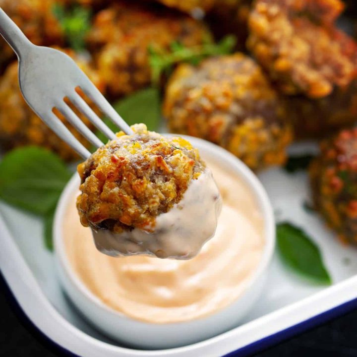 Close up of a golden, delicious sausage ball on a fork dipped in honey maple dijon dipping sauce with a tray of sausage balls in the background