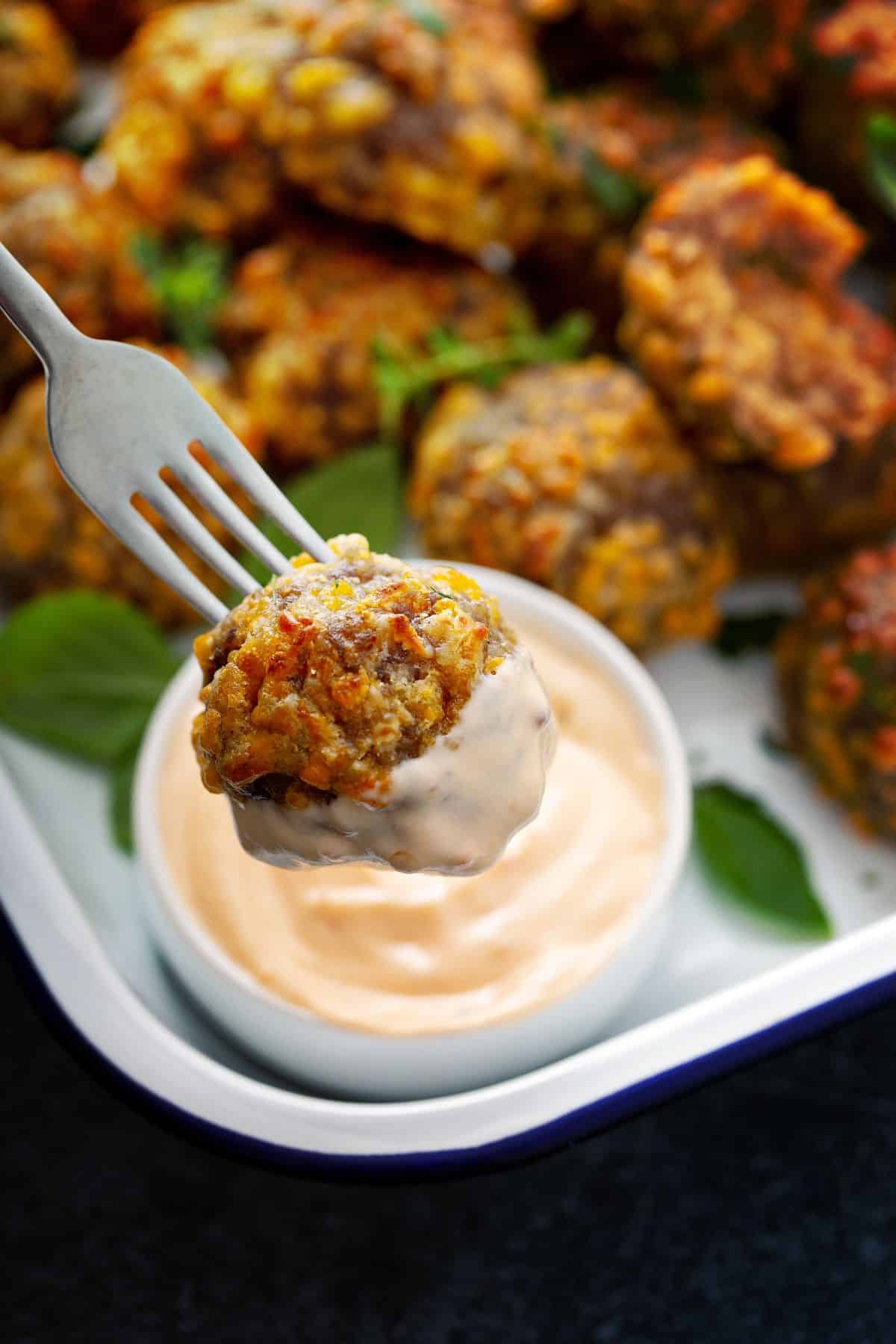 Close up of a golden, crispy sausage ball on a fork, dipped in yummy sauce, with a tray full of sausage balls in the background.