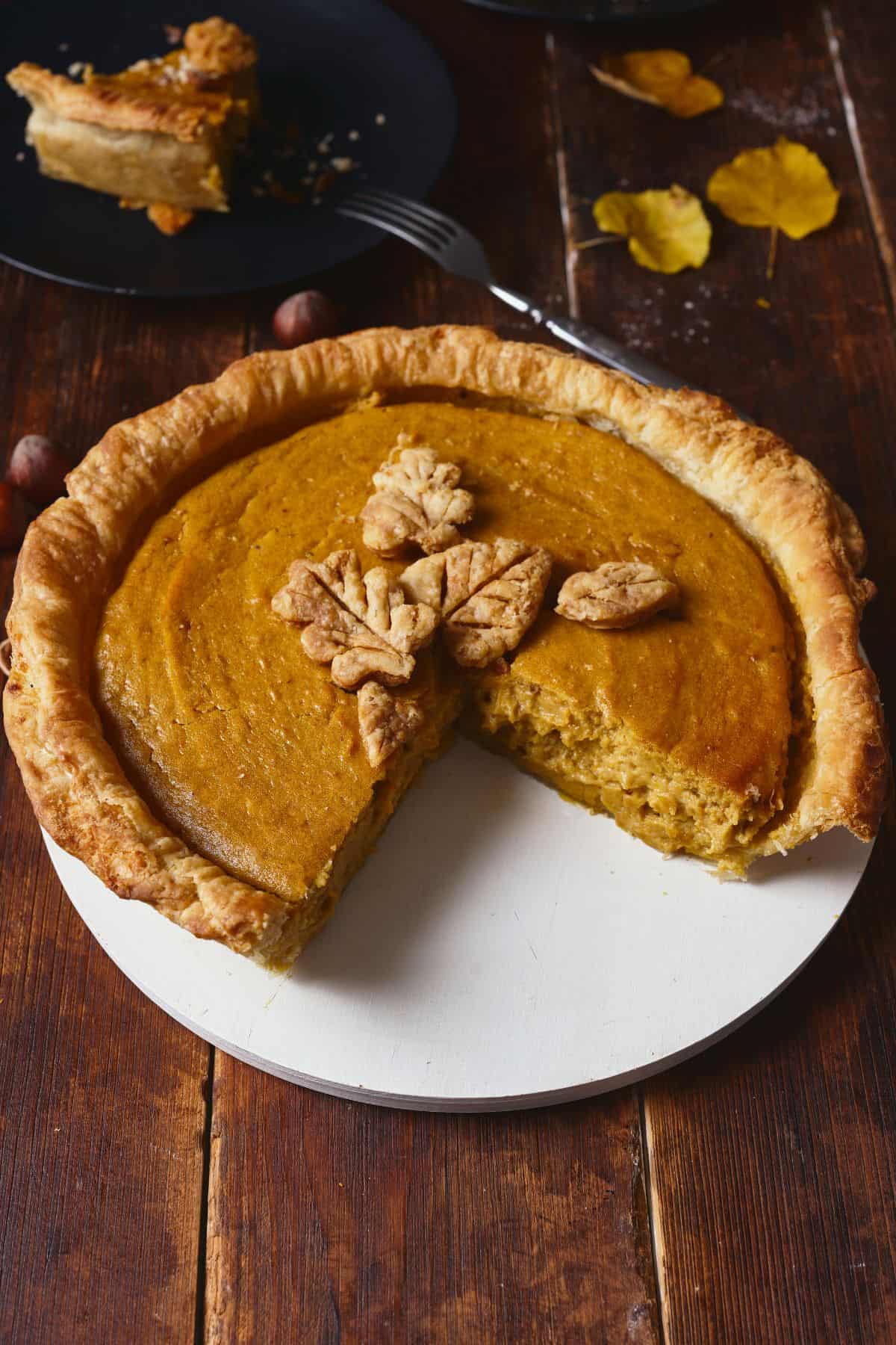 A beautiful, rustic, from-scratch pumpkin pie with homemade crust and leaves on top, sitting beside a cut piece of the pie  on a black plate.