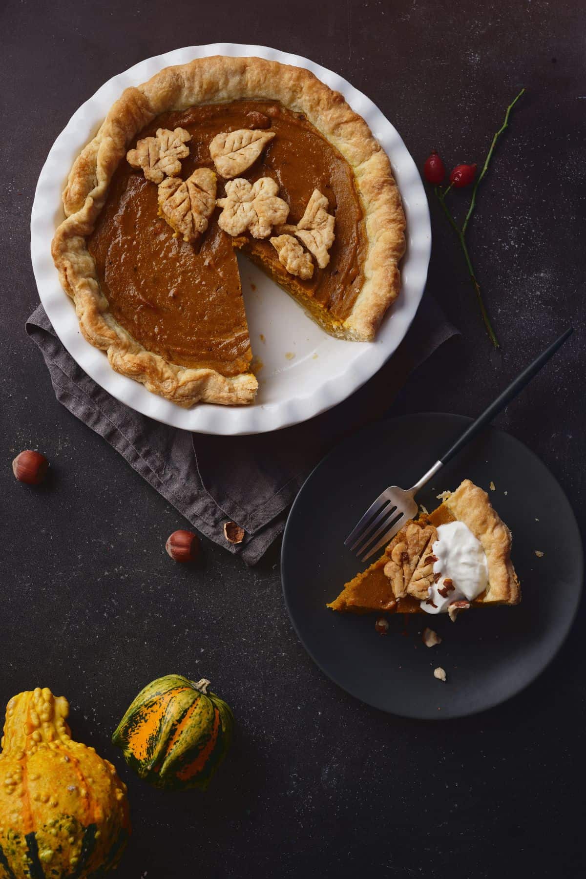 A beautiful, rustic, from-scratch pumpkin pie with homemade crust and leaves on top, sitting beside a cut piece of the pie topped with whipped cream on a black plate.