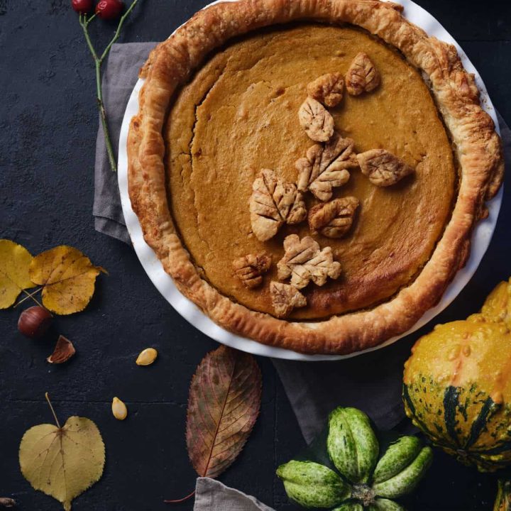 A beautiful, rustic, from-scratch pumpkin pie with homemade crust and leaves on top, sitting beside fresh squash, leaves, and nuts and seeds.