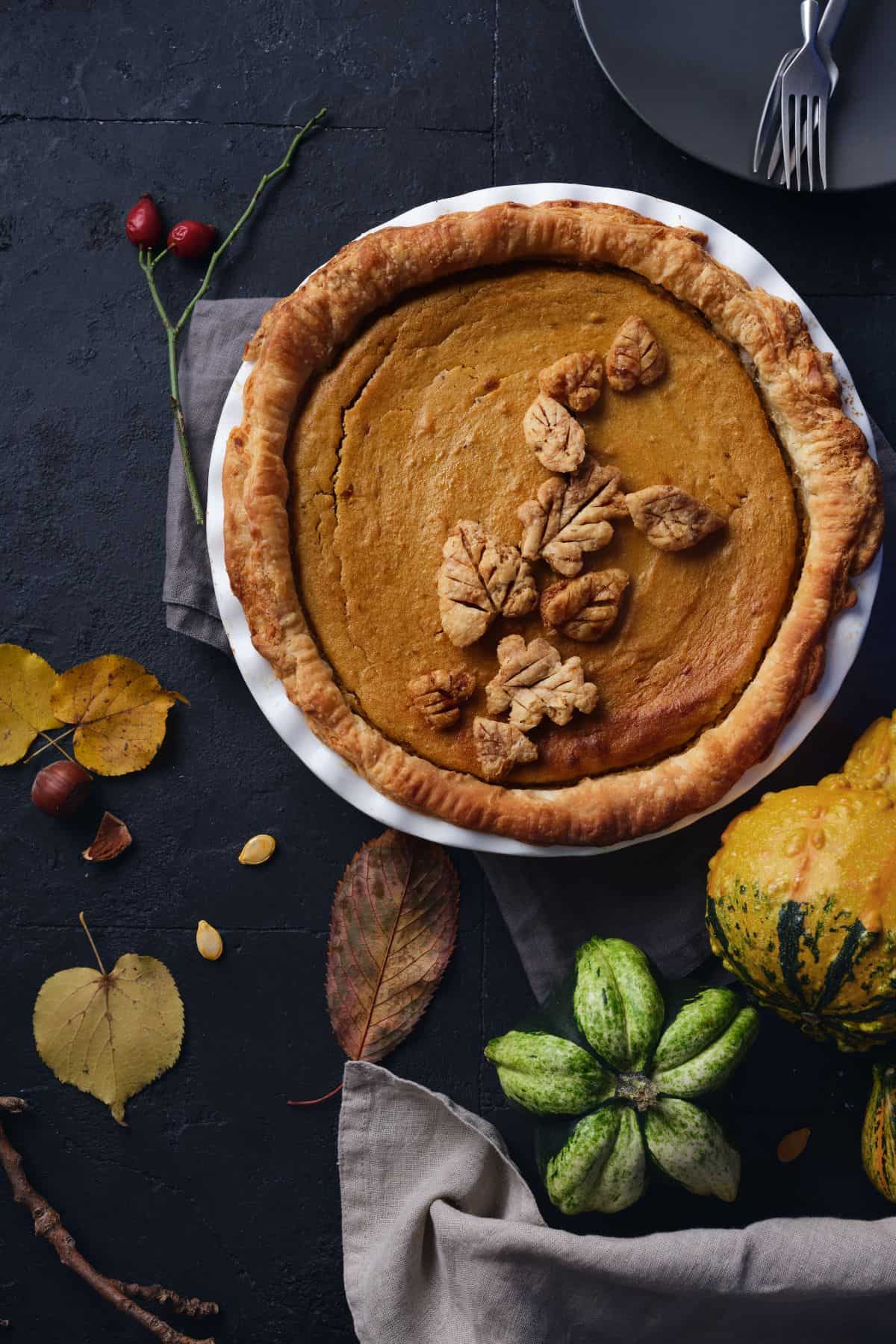 A beautiful, rustic, from-scratch pumpkin pie with homemade crust and leaves on top, sitting beside fresh squash, leaves, and nuts and seeds.