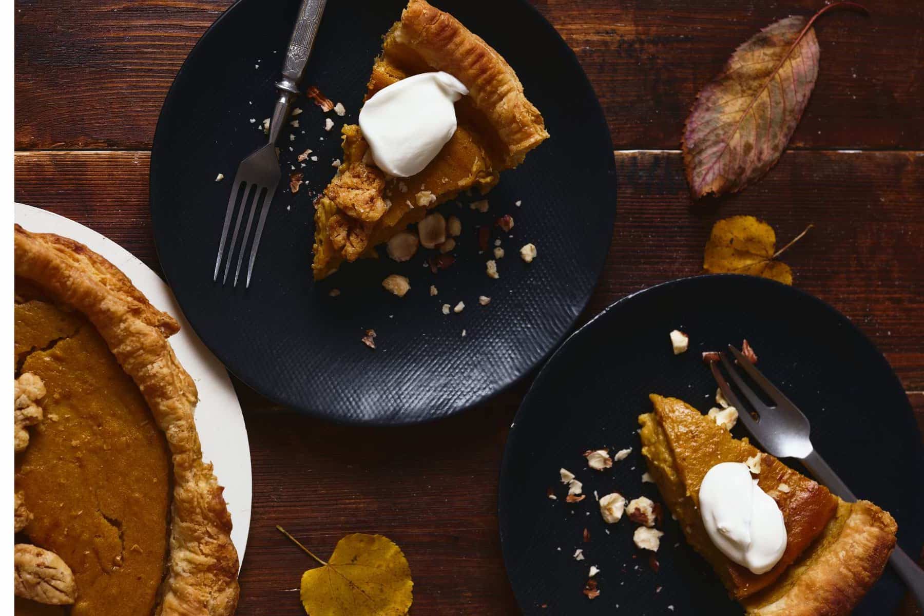 Two black plate with fresh, homemade pumpkin pie topped with whipped cream on a wooden table, with the whole pie in the bottom left of the frame.