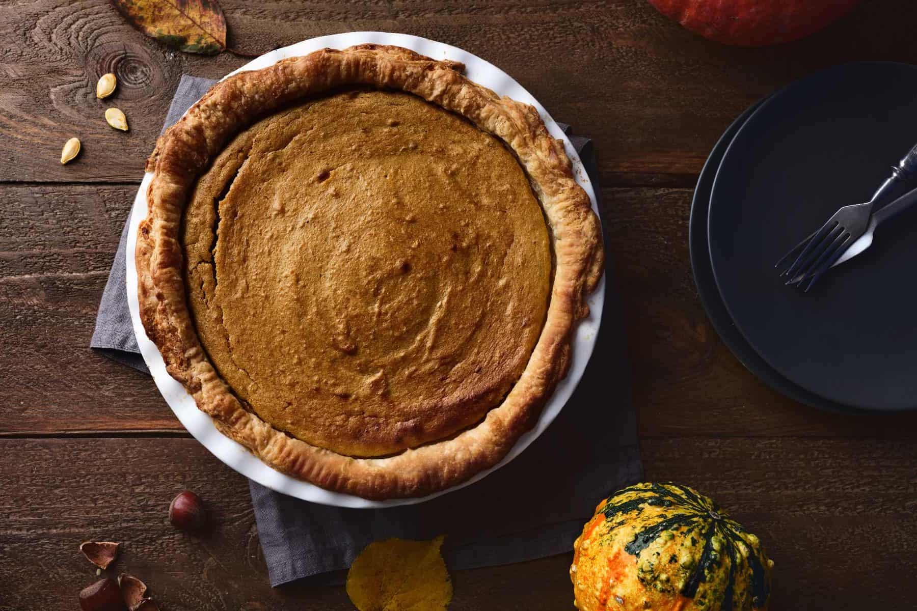 A beautiful, warm, homemade pumpkin pie made from scratch sitting beside black plates, a small squash and pumpkin seeds.