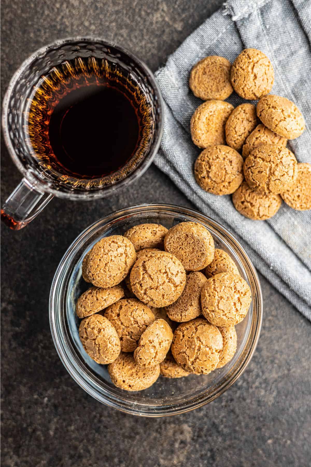 A freshly baked batch of almond sugar cookies - some in a glass bowl and some on a blue linen towel - beside a clear glass mug of black coffee