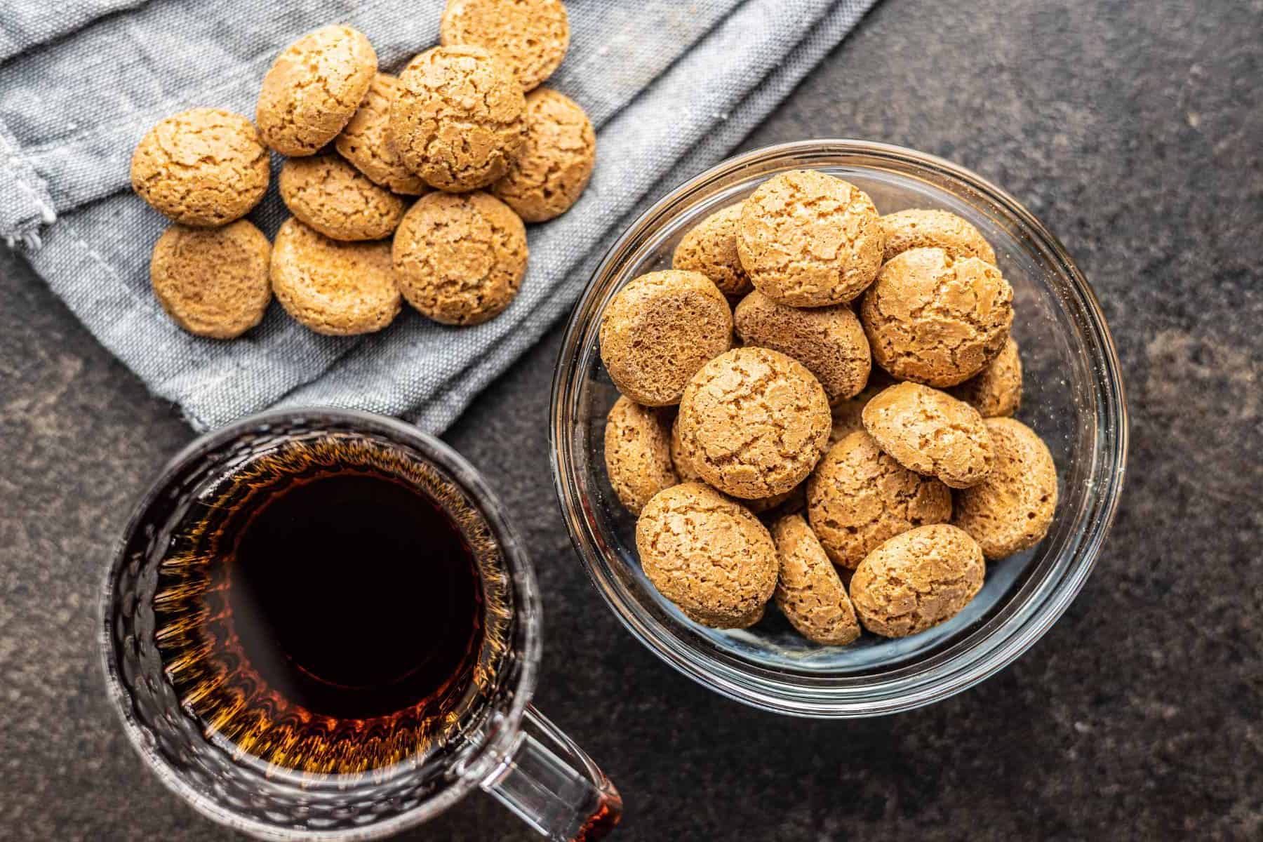 A freshly baked batch of almond sugar cookies - some in a glass bowl and some on a blue linen towel - beside a clear glass mug of black coffee