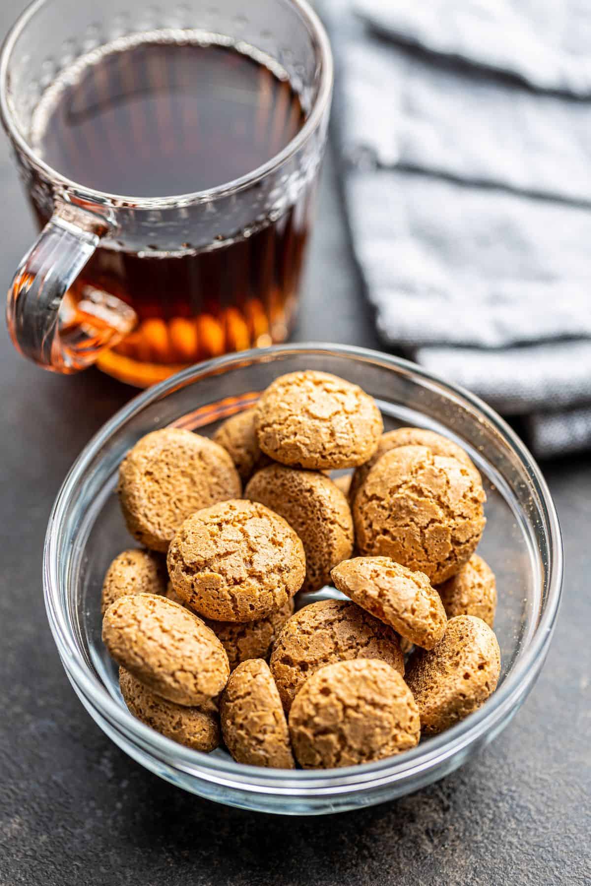 A freshly baked batch of almond sugar cookies in a glass bowl beside a clear glass mug of black coffee and blue linen towel.