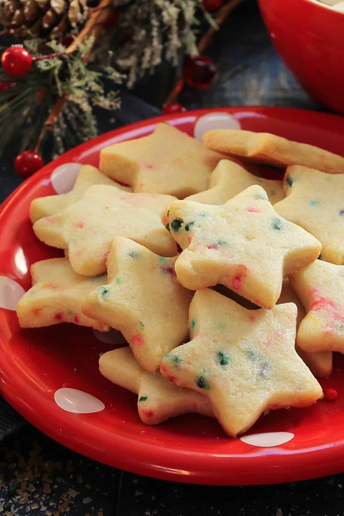 Close up of soft-batch, chewy sprinkle sugar cookies cut into stars with red and green sprinkles sitting on a red plate with greenery and holly in the background.