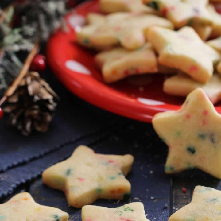 A red plate full of soft-batch, chewy sprinkle sugar cookies cut into stars sitting on a wooden table with greenery, holly, and a few cookies on the table -- a cozy holiday scene!