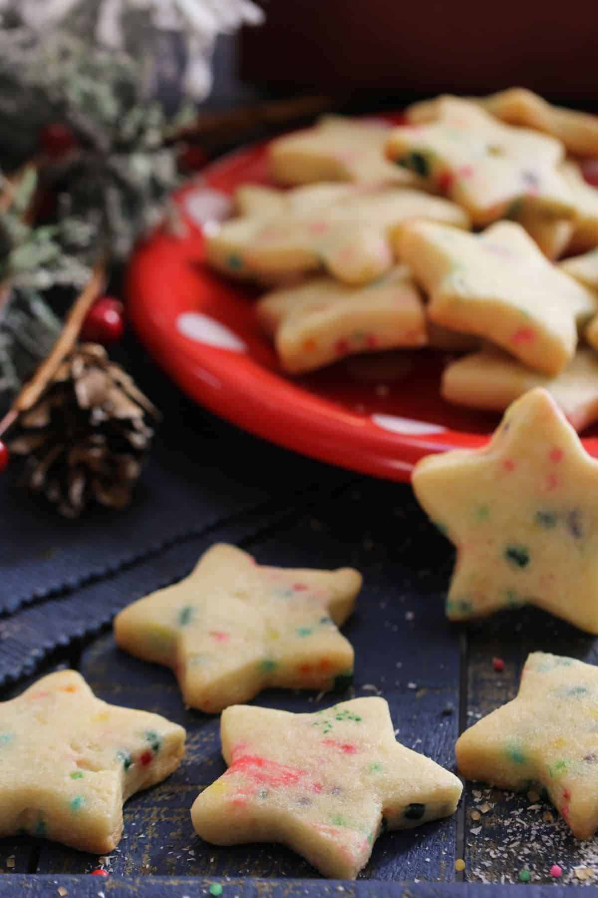 Close up of soft-batch, chewy sprinkle sugar cookies cut into stars with red and green sprinkles sitting on a wooden table with a red plat full of more cookies in the background.